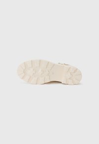 Semelle en caoutchouc avec une surface texturée, de couleur crème clair. Comprend des motifs de crampons et un logo de marque visible.