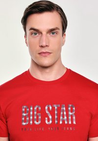Červené bavlněné tričko s velkým textem "BIG STAR" v kostkovaném vzoru, pod nímž je fráze "TVŮJ ŽIVOT. TVÉ DŽÍNY."