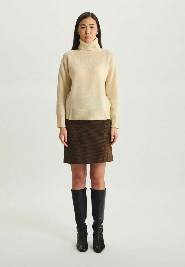 Pull à col roulé crème avec ourlet côtelé, associé à une jupe marron texturée. Le modèle porte des bottes noires élégantes montantes jusqu'aux genoux.
