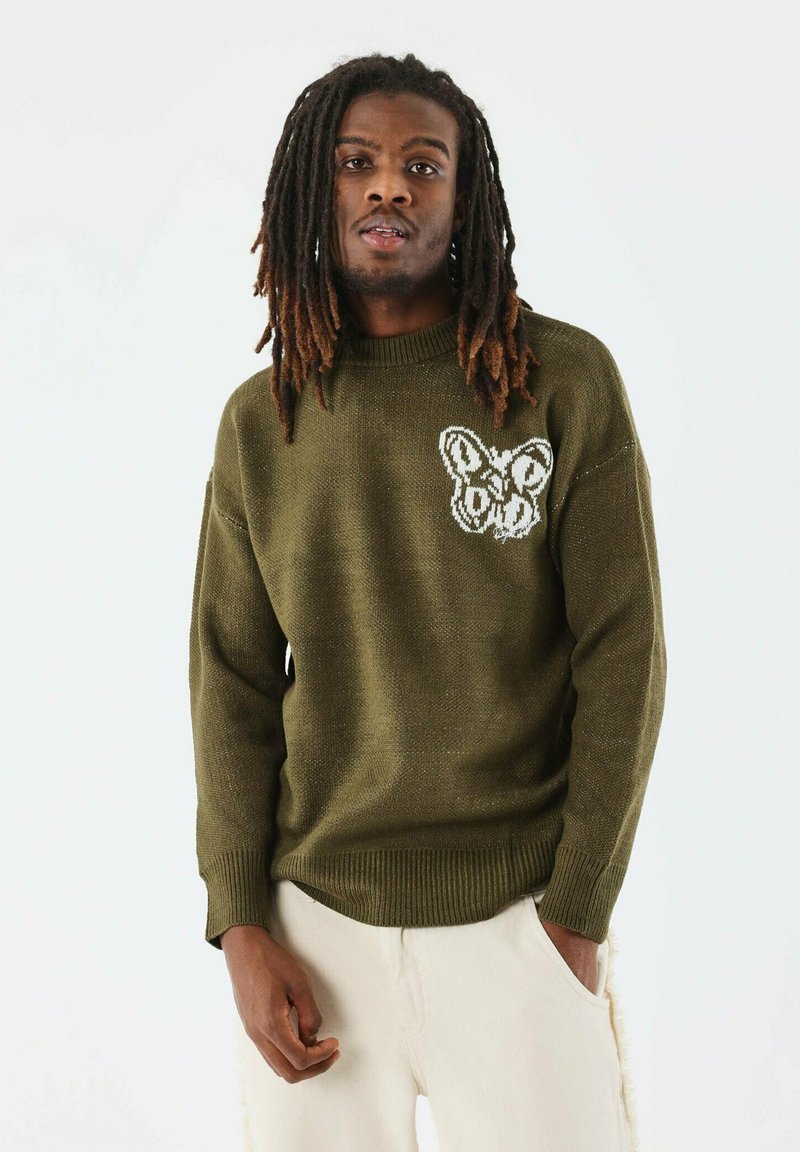 Pull en tricot vert olive avec un col rond, tissu texturé et un motif de papillon blanc sur le côté gauche de la poitrine. Présenté avec un pantalon clair.
