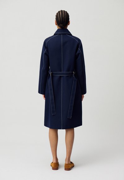 Manteau bleu marine avec une taille ceinturée, manches longues et coutures blanches contrastantes. Il comporte un col et tombe juste en dessous des genoux.