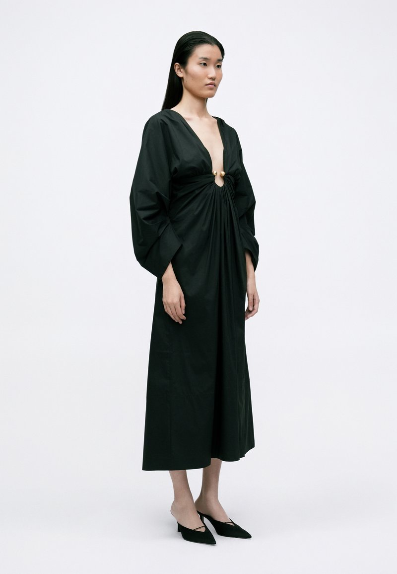 Abito avvolgente nero con scollatura a V profonda, vita arricciata e dettagli in hardware dorato. Presenta maniche lunghe e morbide e una silhouette fluida.