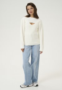 Woman standing wearing a white knit sweater with cutout, light blue wide-leg jeans, and white sneakers on a plain background.