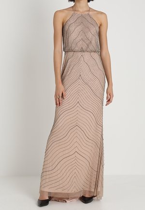 Kobieta w bezrękawowej, długiej do ziemi beżowej sukni z wysokim dekoltem i czarnymi falistymi wzorami, w czarnych butach.