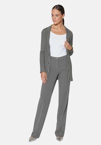 Grijze geribbelde cardigan met knopen, gedragen over een witte top en grijze snitbroek. De outfit omvat zilveren puntige schoenen.
