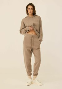 Beżowy skrócony hoodie z logo z przodu, w połączeniu z pasującymi dresami z elastycznym pasem i ściągaczami na kostkach, noszony z białymi trampkami.
