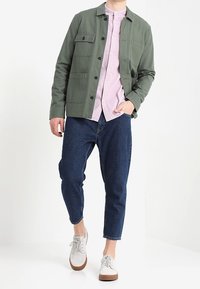 Olivgrüne Jacke mit Taschen über einem hellrosa Hemd mit Knöpfen, kombiniert mit dunkelblauen, schmal zulaufenden Jeans und grauen Sneakers.