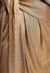 Metallisk guldtyg med ribbad textur, prydnad med veck och en tvistad knutdetalj, vilket skapar visuell och taktil intresse.