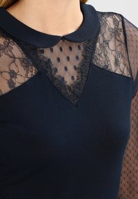 Granatowa bluzka z koronkowym dekoltem ozdobionym kropkami, kołnierzykiem i teksturowanymi rękawami, łącząca gładkie i przezroczyste materiały.