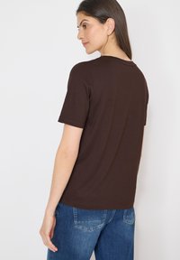 Femme portant un t-shirt marron à manches courtes et un jean bleu, légèrement tournée pour montrer le dos du t-shirt sur un fond blanc.