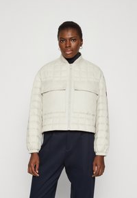 Quilted lettvekts puffer-jakke i lys beige med elastiske mansjetter, glidelås foran og to brystlommer. Minimalistisk design og cropped lengde.