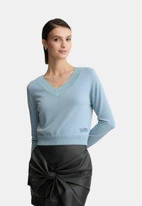 Maglione blu a V con orlo a coste e dettagli scintillanti intorno allo scollo, abbinato a una gonna nera con un grande dettaglio a fiocco.