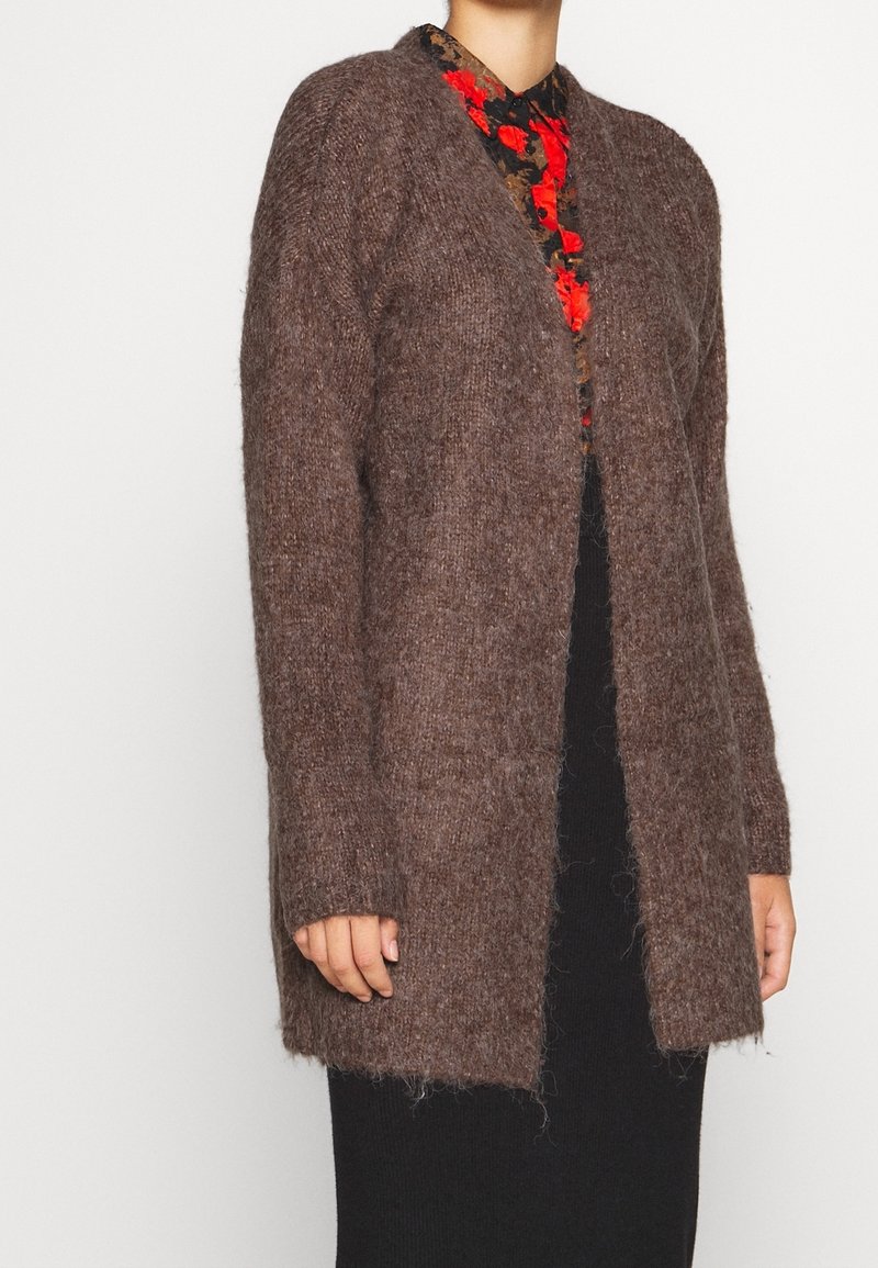 Cardigan marron texturé à manches longues, devant ouvert, en matière duveteuse, porté par-dessus une robe noire ajustée. Motif floral rouge visible en dessous.