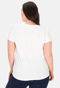 Hvid t-shirt med korte ærmer, rund halsudskæring og opkrøllede ærmer, der har en glat tekstur og et simpelt design. Bæres med mørkeblå jeans.