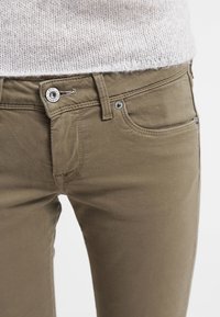 Olivgröna bomullsbukser med knäppning, fem fickor och en texturerad midja. Sömmen är synlig överallt.