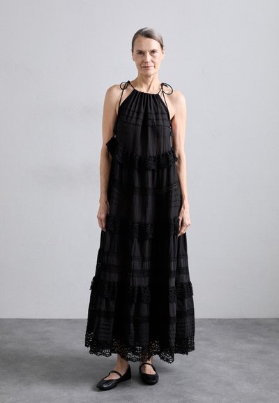 Vestido largo negro con tejido texturizado, que presenta volantes en capas y acentos de encaje. Combinado con zapatos planos negros.