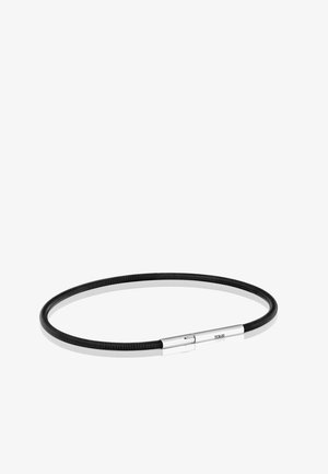 Collar flexible negro enroscado con un cierre magnético plateado pulido grabado con "TOUS" sobre un fondo blanco.