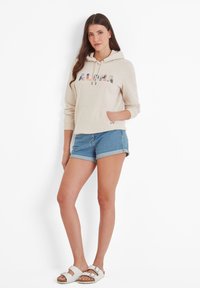 Beige hoodie with "ALOHA" in colorful floral letters, front pocket, paired with rolled denim shorts, and white slide sandals.