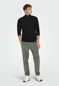 Svart zip-neck tröja, figurnära design, bärs med grå kordflossbyxor och krämfärgade sneakers med gröna detaljer. Slät stickad struktur.