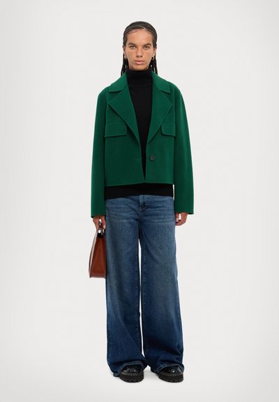 Green cropped jacket with two front pockets, black turtleneck, wide-leg blue jeans, and black chunky shoes, holding a brown handbag.