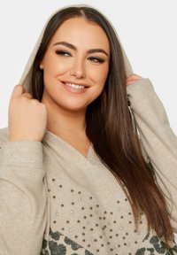Beige hoodie met een v-hals ontwerp, versierd met donkere en lichte studs, met een accent van luipaardprint en een zachte, gestructureerde stof.