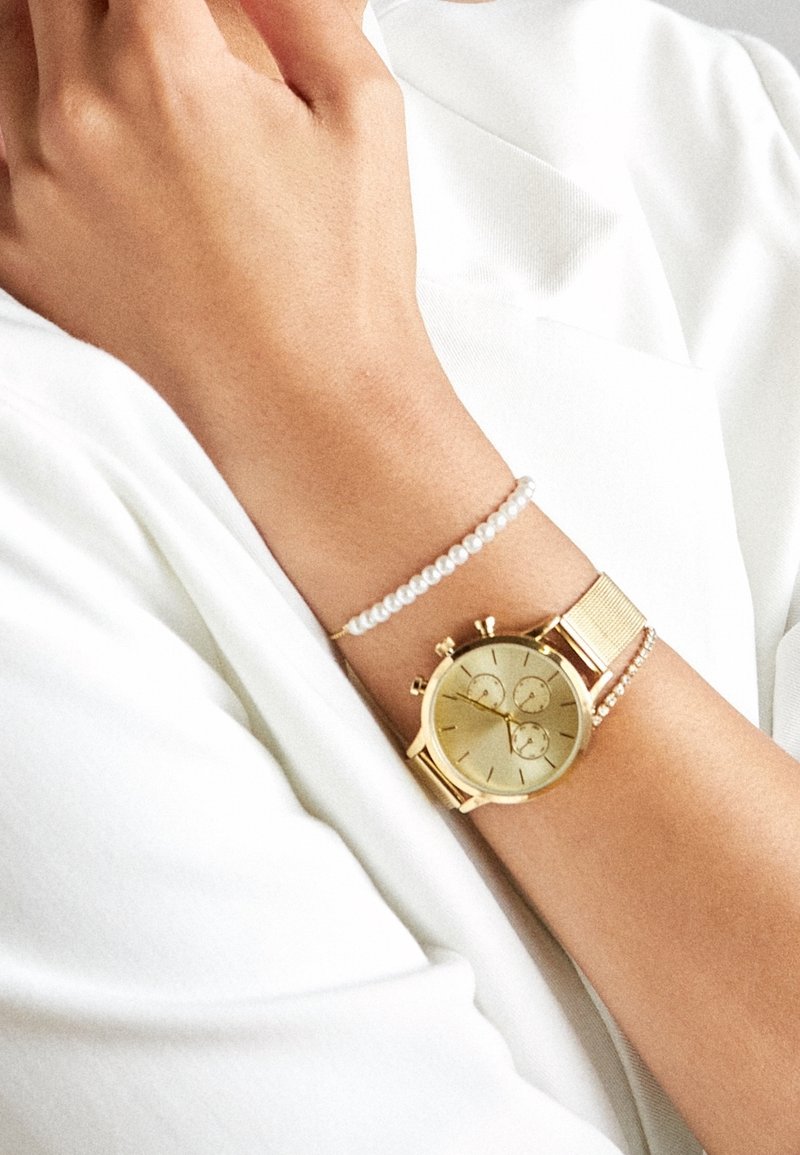 Poignet avec montre chronographe en or et deux bracelets délicats en perles sur fond de vêtements blancs.