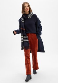 Granatowy sweter, kraciasty szalik w czarno-białe paski, rdzawy kordowy spodnie, czarne botki na obcasie, casualowa warstwowa stylizacja.