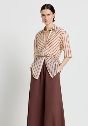 Donna con camicia beige a righe ruggine con bottoni e vita annodata, pantaloni marroni a gamba larga, in piedi con le mani nelle tasche su sfondo bianco.