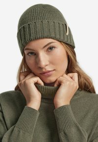 Olivgrön stickad mössa med ribbad design och en läderdetaljtagg. Bärs med en matchande högkragad tröja. Mjuk textur, avslappnad passform.