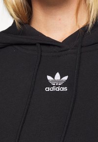 Černá mikina s kapucí vyrobená z bavlněné směsi. Má bílé vyšívané logo Adidas s třemi pruhy níže.