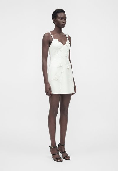 Γυναίκα στέκεται φορώντας λευκό μίνι φόρεμα με λουλουδένιες εφαρμογές και μαύρα ψηλοτάκουνα σανδάλια σε απλό φωτεινό φόντο.