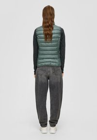 Ljusgrön vadderad väst med horisontella quiltade sektioner, tillsammans med löst sittande grå denimjeans och vita sneakers. Platt bakgrund.