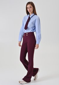 Femme portant une chemise bleue boutonnée, une cravate bordeaux, un pantalon taille haute bordeaux avec des boutons dorés, et des baskets blanches, debout devant un fond uni.