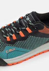 Zapatilla deportiva negra y turquesa con material de malla texturizada, detalles en naranja y una suela gris con tracción multidireccional.