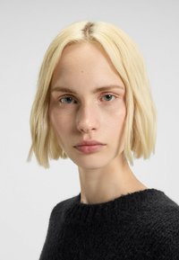 Capelli biondi, taglio a caschetto corto; pelle liscia, trucco minimal. Maglione nero in tessuto a maglia, con scollatura rotonda; sfondo neutro.