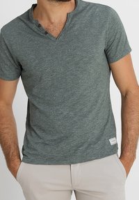 Mężczyzna w szarym T-shircie z krótkim rękawem i dekoltem w serek, zapinanym na trzy guziki, w beżowych spodniach, z rękami w kieszeniach, na jednolitym tle.