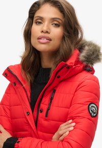 Giacca puffer rossa con tasche con zip, cappuccio con bordatura di pelliccia e dettagli neri. Presenta un design trapuntato e una patch con il marchio sulla manica.