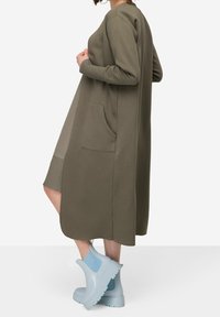 Largo vestido verde oliva con un dobladillo redondeado, mangas largas y bolsillos laterales. Lleva unas botas de tobillo azul claro con una suela gruesa.