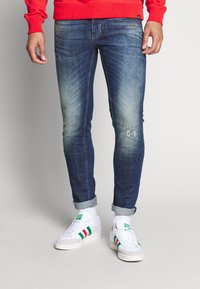 Upravené modré džínové kalhoty s lehkým zesvětlením a zaprášením, ohrnuté nohavice, v kombinaci s bílými teniskami se zelenými a červenými pruhy.