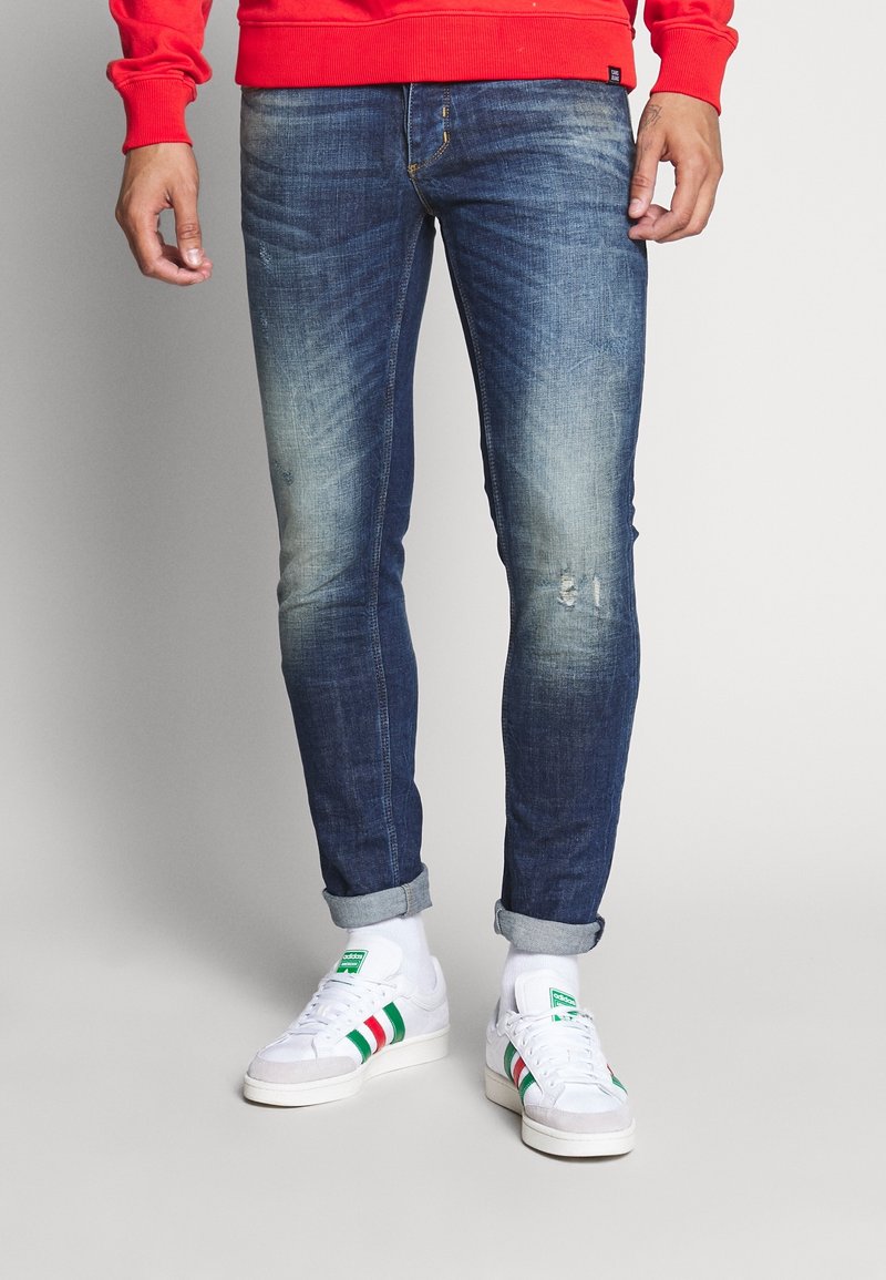 Upravené modré džínové kalhoty s lehkým zesvětlením a zaprášením, ohrnuté nohavice, v kombinaci s bílými teniskami se zelenými a červenými pruhy.