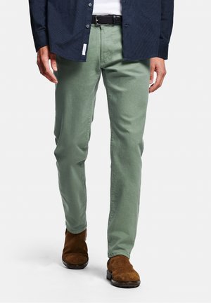 Homme portant un pantalon droit vert sauge, des bottes en daim marron, une ceinture noire, une chemise blanche et une chemise à boutons bleu marine, debout devant un fond blanc.