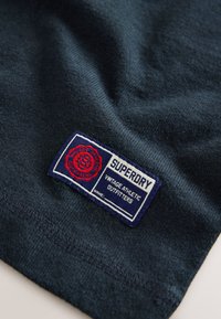 Mörkblått bomullstyg från T-shirt med en fastsydd vävd etikett som visar "SUPERDRY" och "VINTAGE ATHLETIC OUTFITTERS" i rött och vitt.