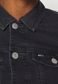 Detailní záběr na límec a horní část hrudi tmavě modré džínsové bundy se stříbrnými knoflíky a malým logem značky na klopě levé kapsy.