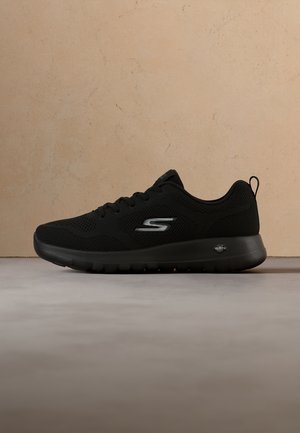 Zwarte Skechers sportschoen met ademend mesh bovenwerk, vetersluiting en grijs "S" logo aan de zijkant, getoond tegen een beige muur en grijze vloer.