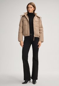 Brązowa pikowana kurtka z geometrycznym wzorem, krótki krój, duży kołnierz i przednie kieszenie. Nosi na sobie czarny golf i czarne dzwony.