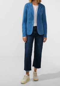 Blazer azul con corte entallado, dos bolsillos frontales, llevado sobre una camisa blanca. Combinado con vaqueros de pierna ancha de mezclilla oscura y zapatillas claras.