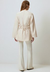 Beige, tailored jacket with a cinched back and long sleeves, paired with white flared pants. Smooth texture, minimalistic design.
