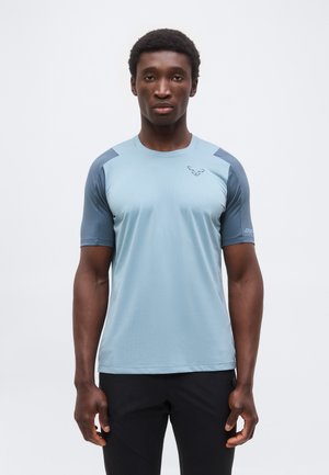 Egy férfi áll, előre néz, világoskék és szürke rövid ujjú sportpólót és fekete nadrágot visel, sima fehér háttér előtt.