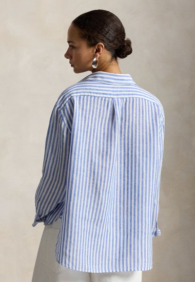 Kvinne med hår i en knute, iført sølvsølvringer og en løs blå og hvit skjorte med vertikale striper, stående mot en ensfarget bakgrunn.