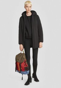 Cappotto imbottito nero con cappuccio, top nero e leggings, con uno zaino multicolore (marrone, rosso, blu) con hardware e cinghie uniche.